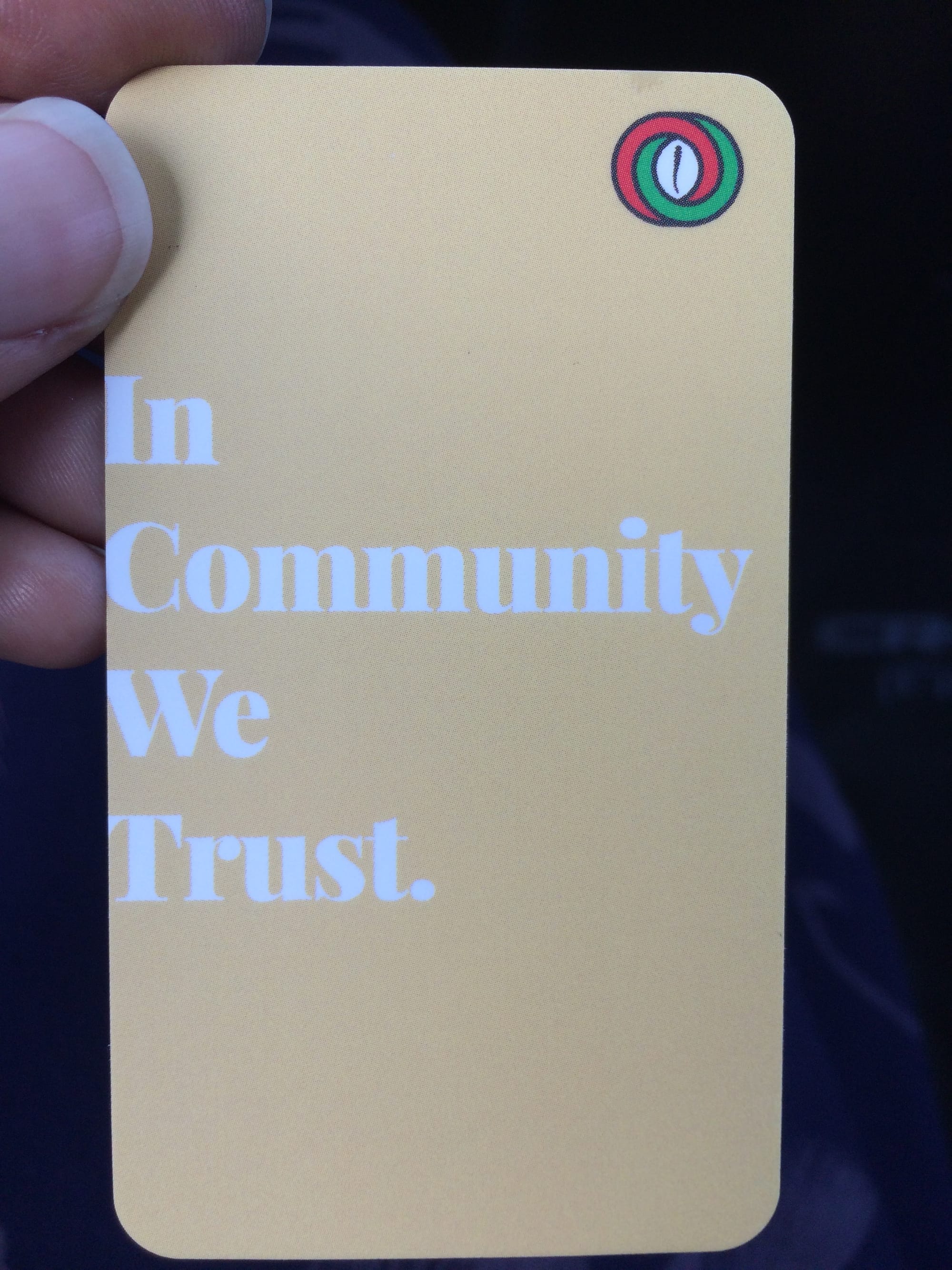 Brown hand with thumbnail showing holds a light yellow business card that says In Community We Trust with a red black and green cowry shell logo in the upper right corner. 